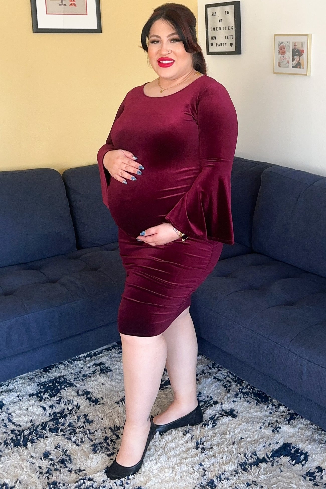 woman wearing a burgundy maternity dress with bell sleeves, showing her baby bump while standing on a couch