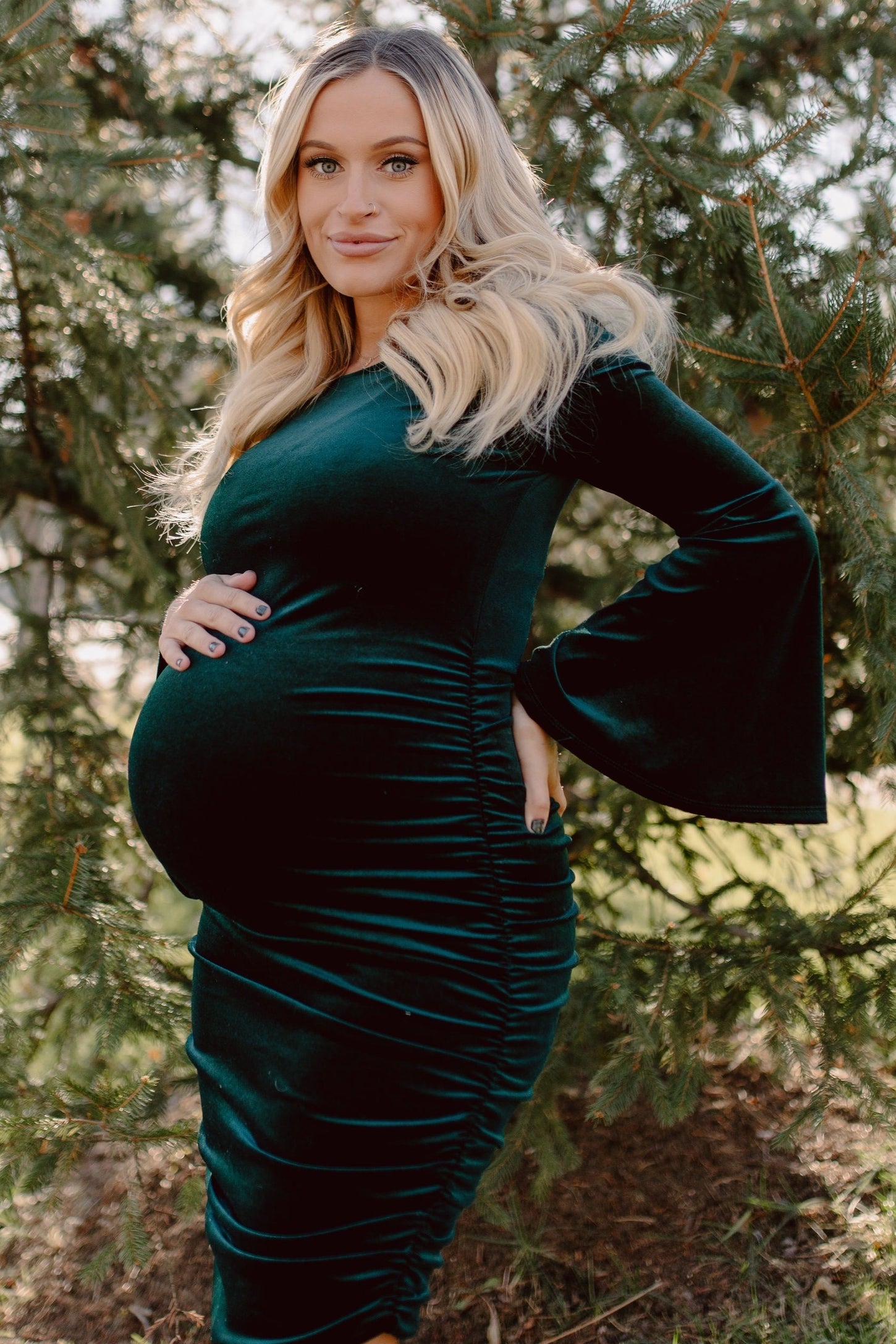 woman wearing a dark green velvet maternity dress with bell sleeves and gathered details, showcasing her baby bump