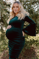 woman wearing a dark green velvet maternity dress with bell sleeves and gathered details, showcasing her baby bump