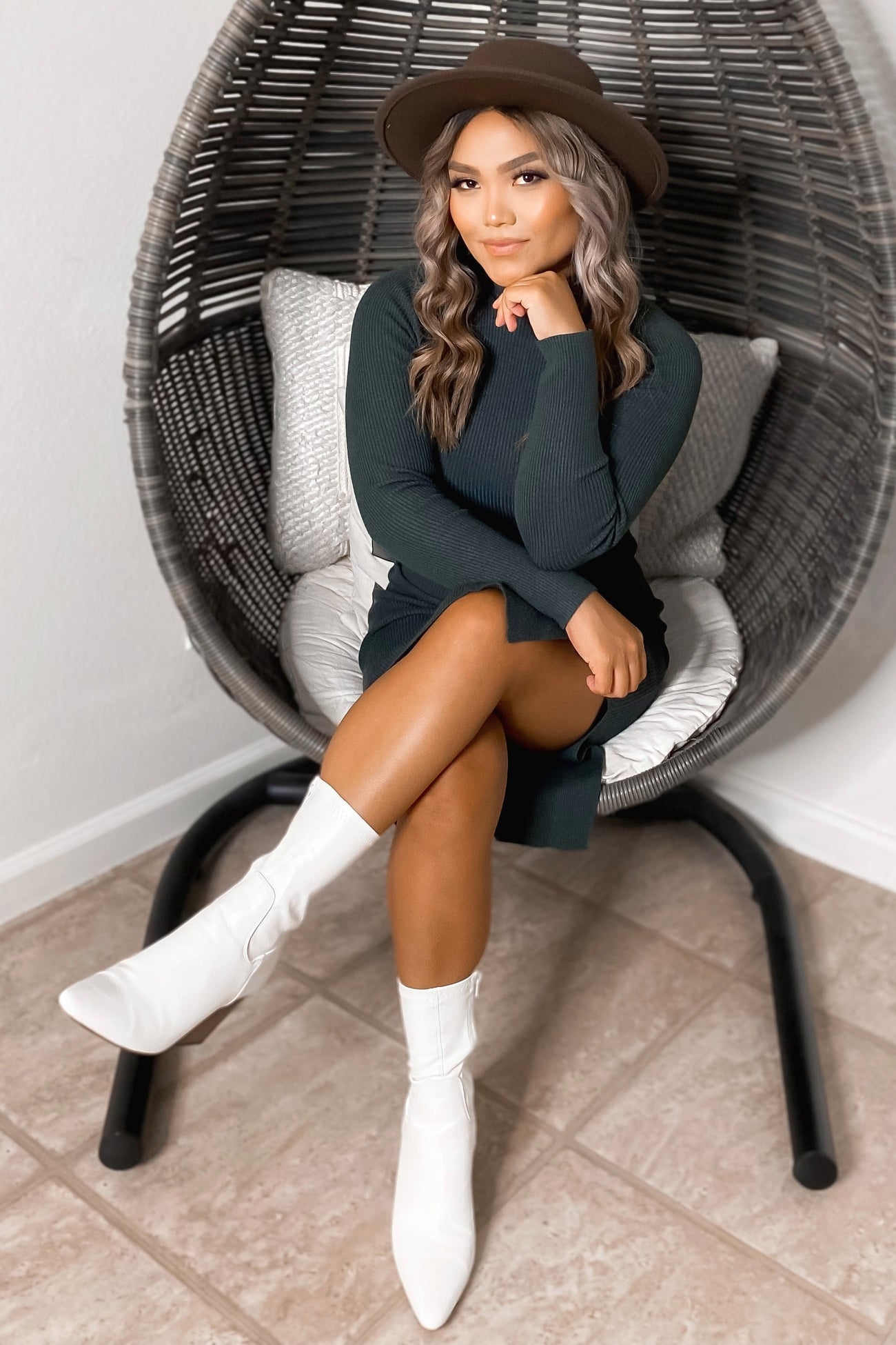 woman wearing a dark green long sleeve sweater dress and white heeled ankle boots, sitting in a hanging chair