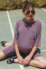 woman wearing a mauve maternity short sleeve romper with a front pocket and a visible baby bump, sitting on the ground