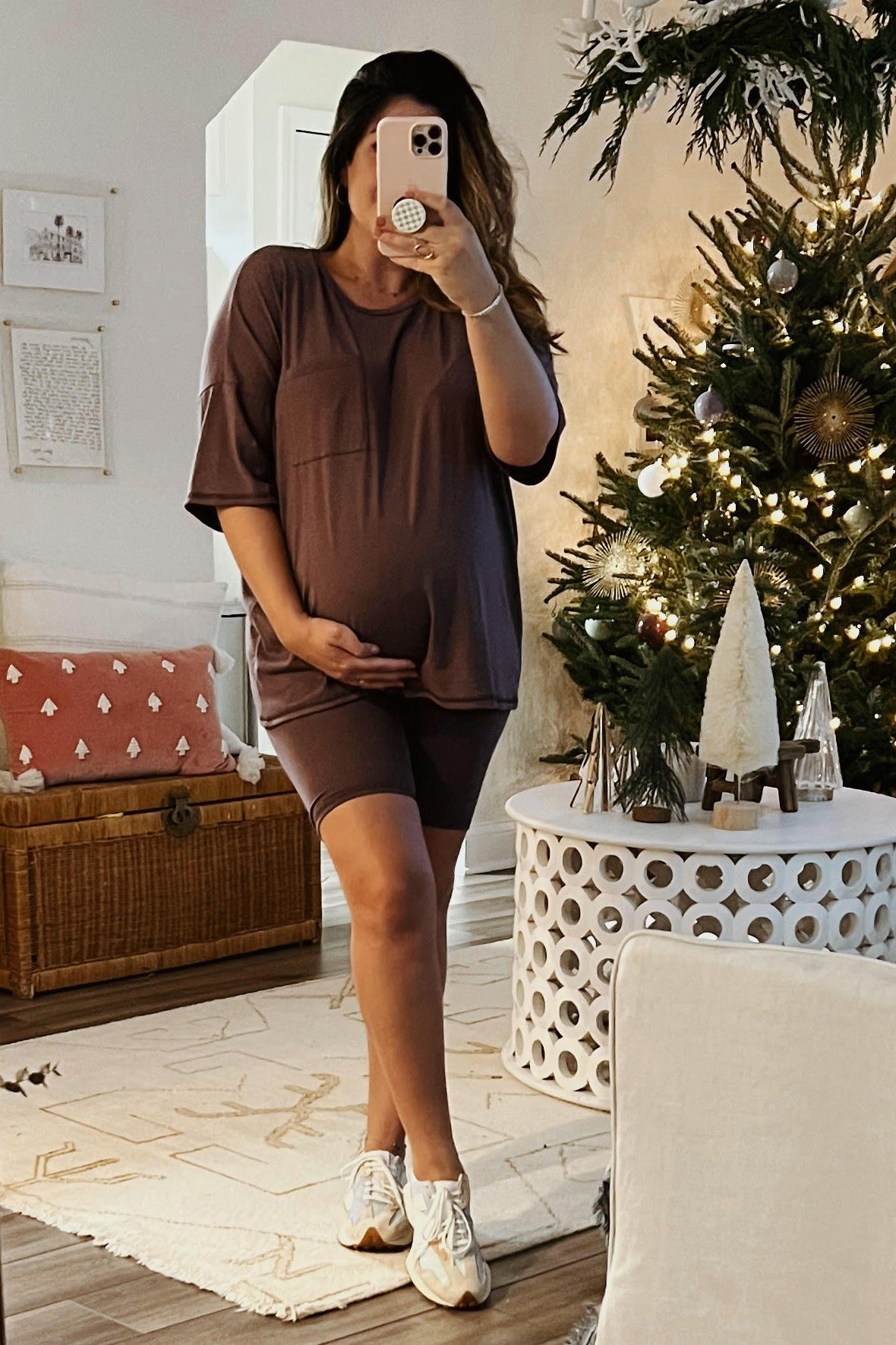 woman wearing a mauve maternity top and bike shorts, holding her baby bump in front of a decorated Christmas tree