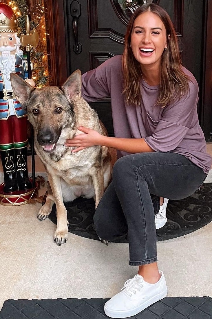 woman wearing a mauve oversized t-shirt and black jeans, sitting on the floor with a dog and smiling