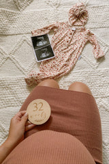 Pregnant woman holding 32 weeks milestone disc next to floral baby gown and ultrasound photos