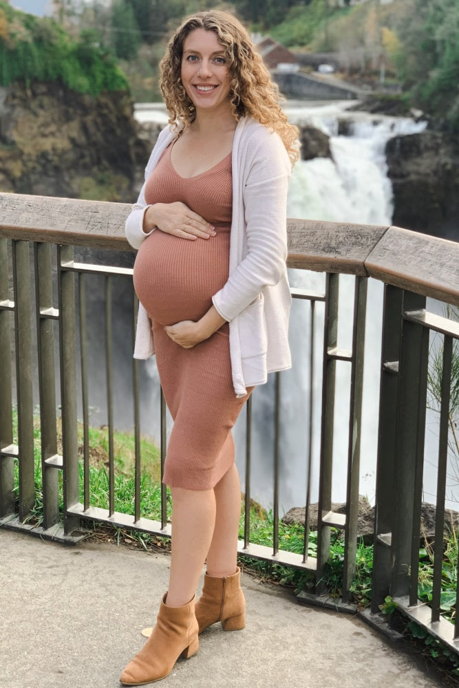 Smiling pregnant woman in a terracotta ribbed dress and white cardigan standing by a waterfall