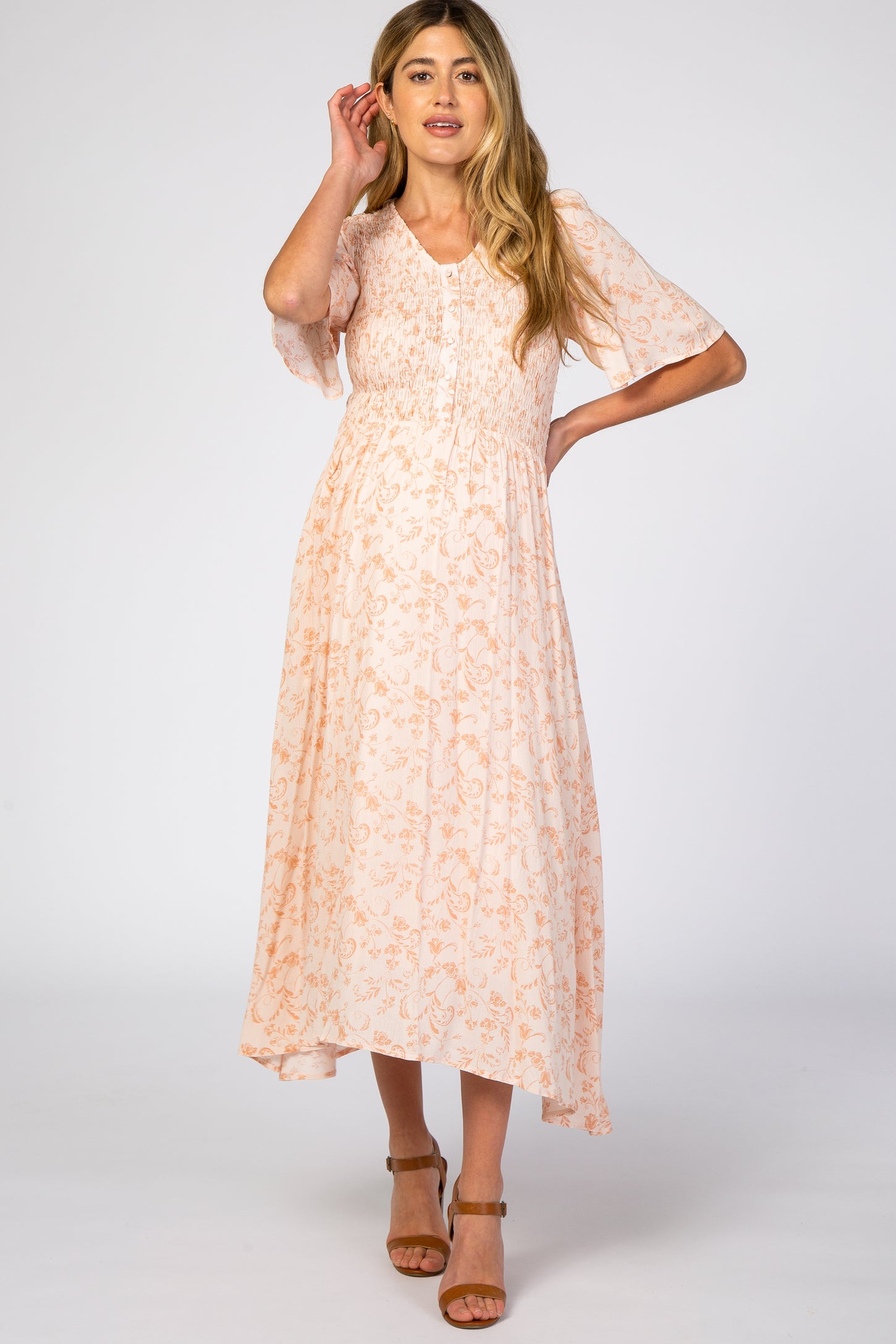woman wearing a light peach floral maternity dress with short sleeves and a smocked bodice, standing with one hand on her hair