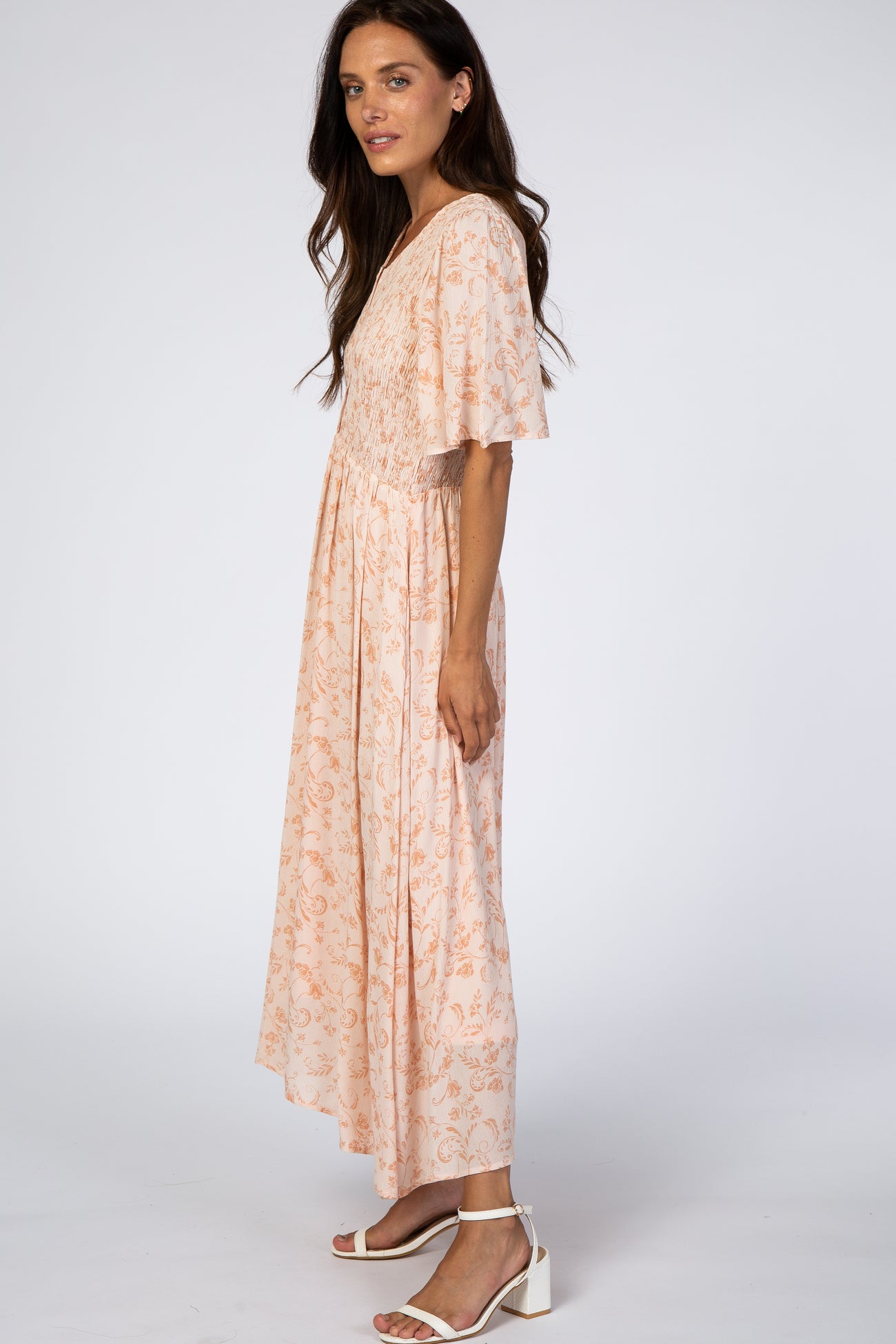 woman wearing a peach floral maxi dress with flutter sleeves and smocked bodice, standing sideways against a white background