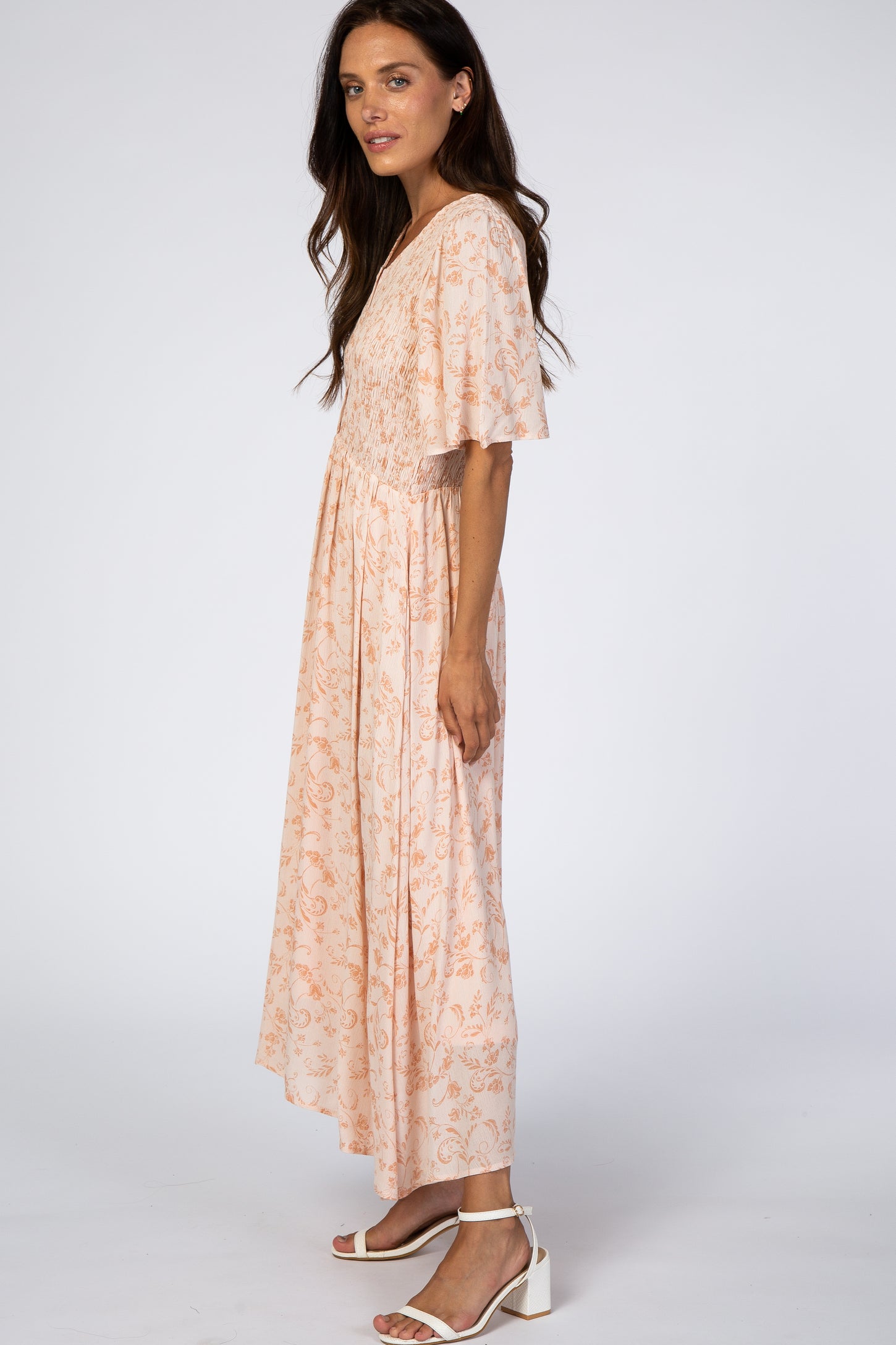 woman wearing a peach floral maxi dress with flutter sleeves and smocked bodice, standing sideways against a white background