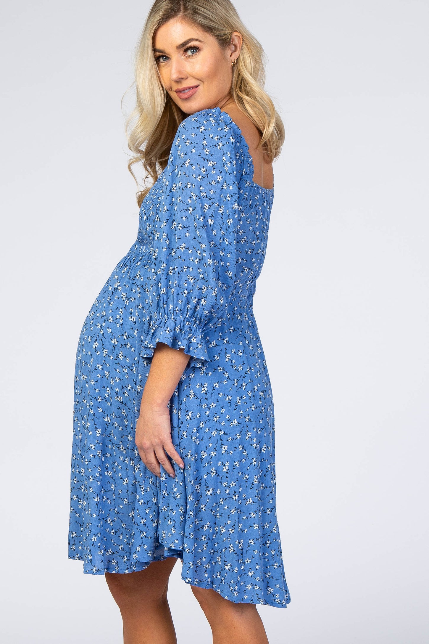 Pregnant woman wearing a blue floral maternity dress with ruffled sleeves against a white background