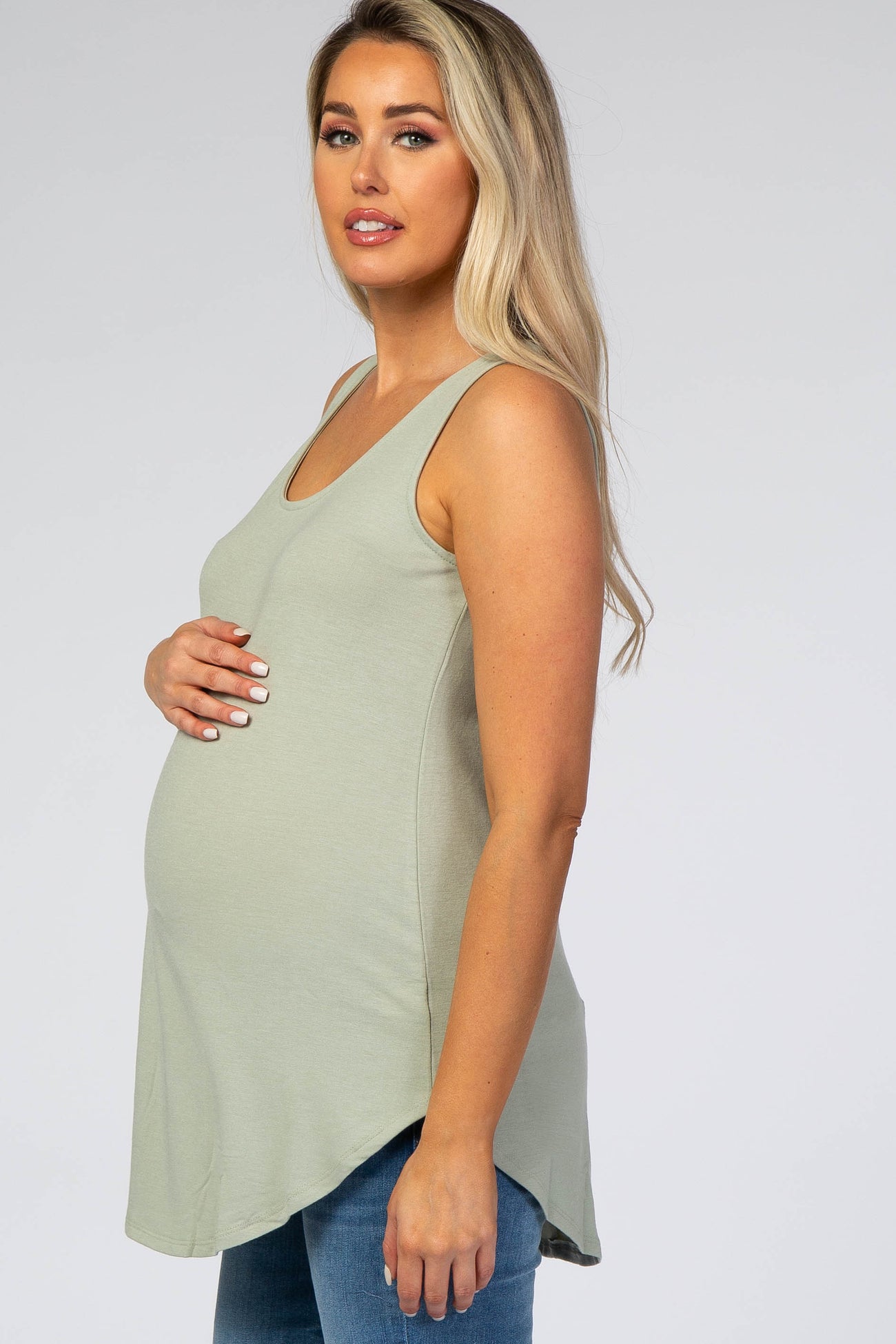 pregnant woman wearing a sage green maternity tank top and blue jeans against a white background