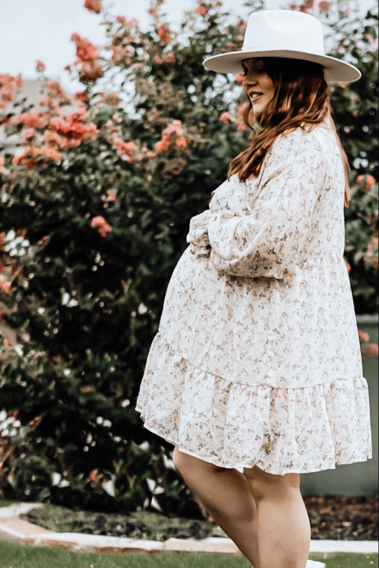 pregnant woman in floral maternity dress and white wide brim hat standing in front of flowering bushes