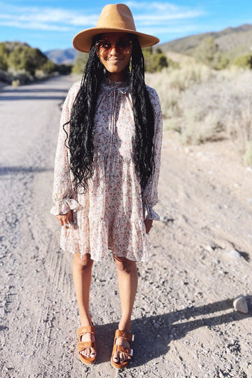 woman wearing a pink floral long sleeve dress with a ruffled hem, brown hat, and brown sandals, standing outdoors