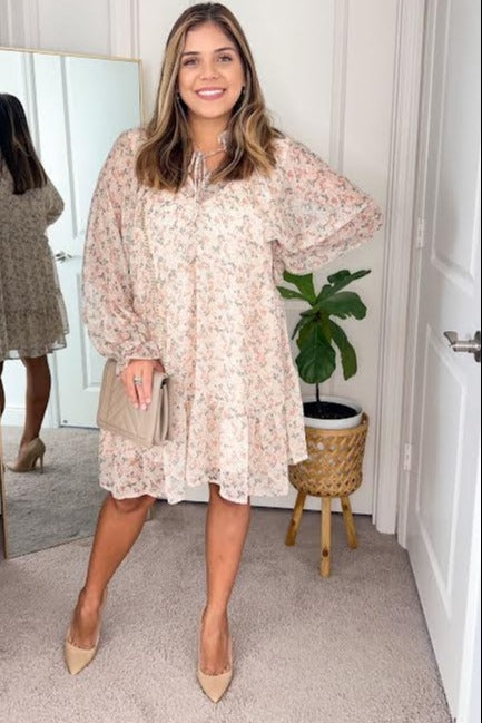 woman wearing a long-sleeved floral mini dress and beige heels holding a quilted clutch bag