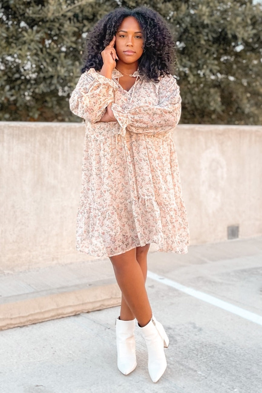 woman wearing a light pink floral mini dress and white ankle boots, standing outdoors with curly hair