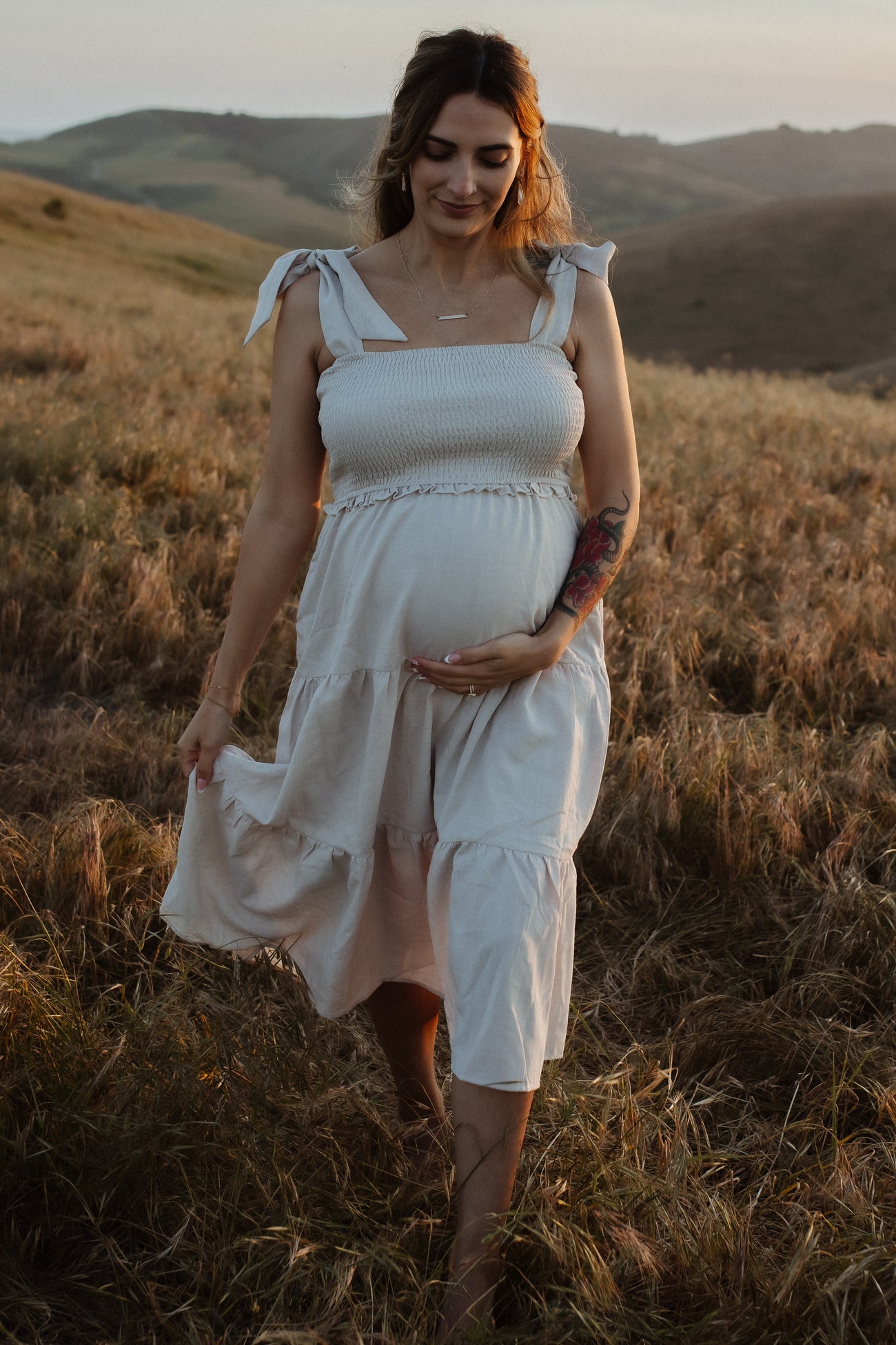 Pregnant woman in a white smocked dress walking through a golden field at sunset with hills in the background