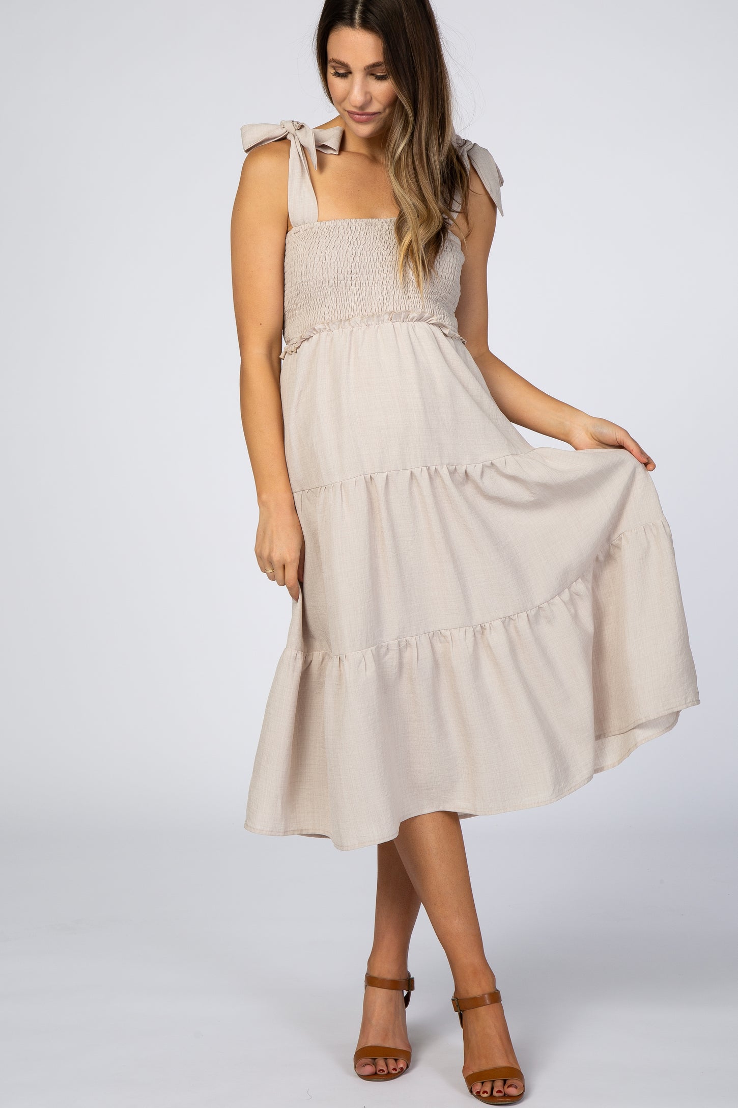 Woman wearing a beige smocked maternity midi dress with tie-straps and a tiered skirt