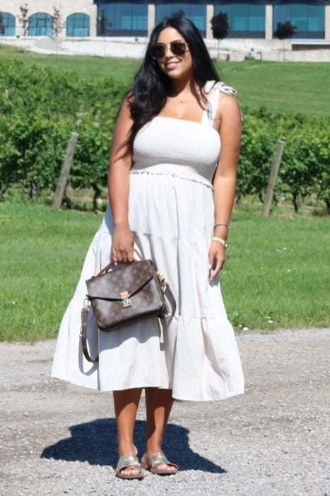 woman wearing a cream-colored tiered maxi dress with adjustable straps, holding a handbag, standing on a grassy area