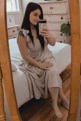 woman wearing a light beige maxi dress with bow straps, seated on a bed and taking a selfie in a mirror