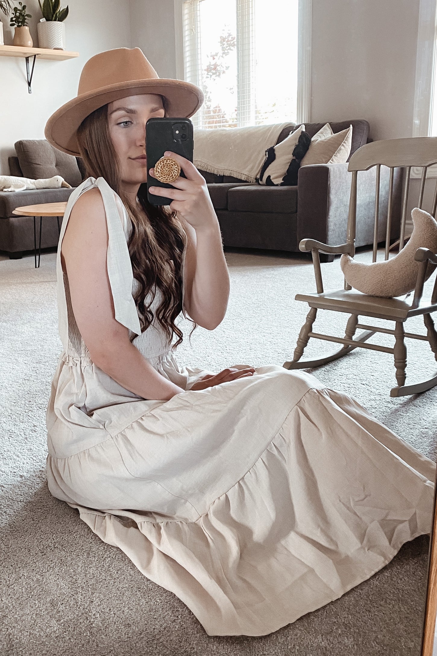 woman sitting on the floor wearing a beige sleeveless maxi dress and a wide-brimmed hat in a cozy living room setting