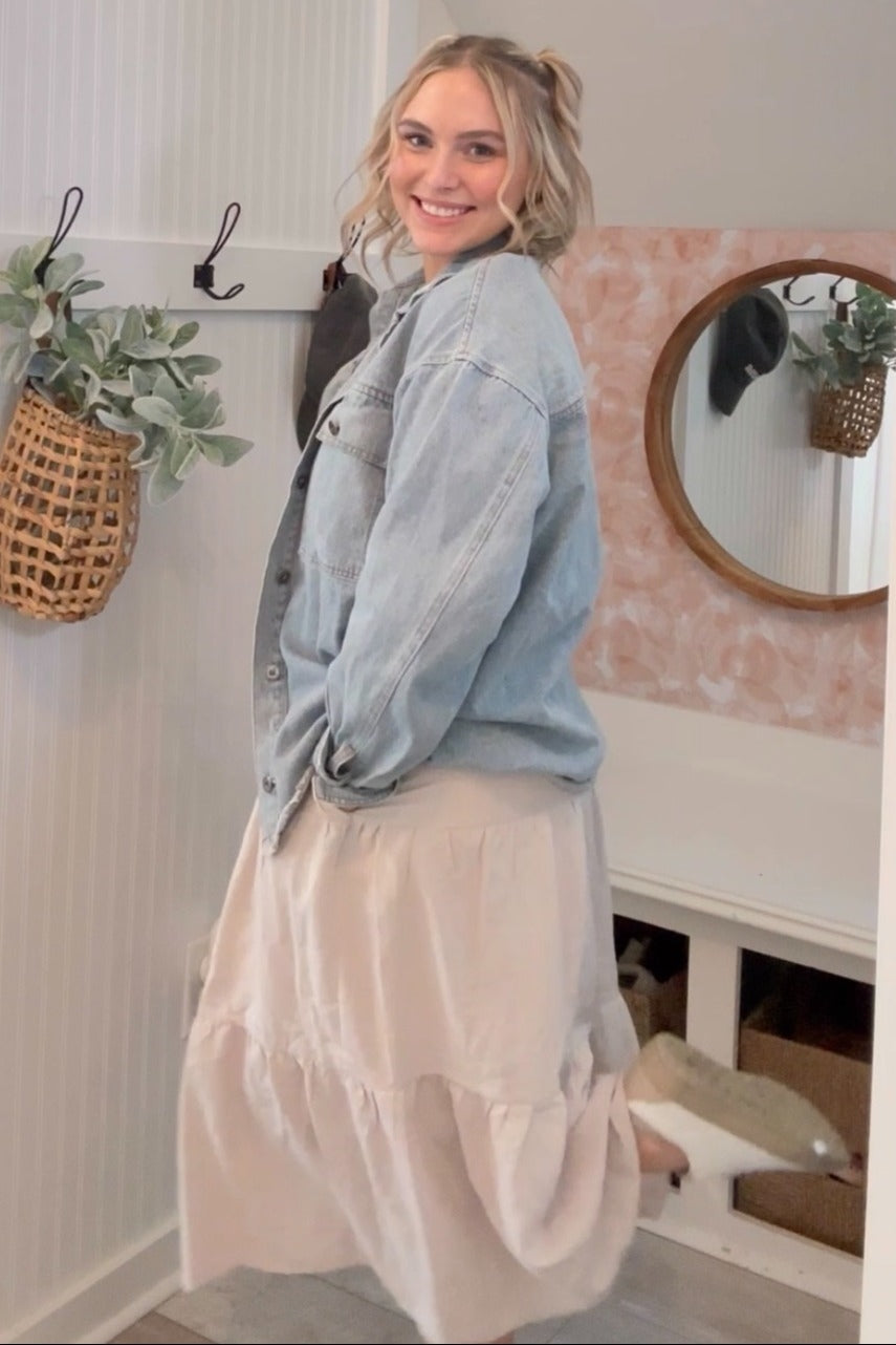 woman wearing a light denim oversized jacket and a flowy beige tiered skirt, smiling and standing in a bright room