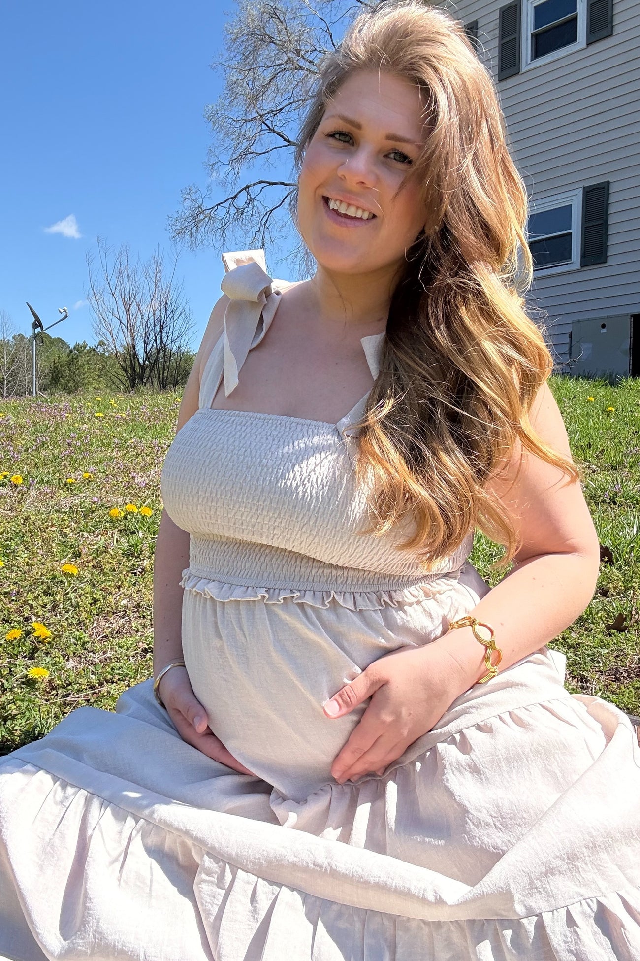 woman wearing a light beige maternity dress with smocked bodice and bow straps, sitting outdoors and holding her baby bump