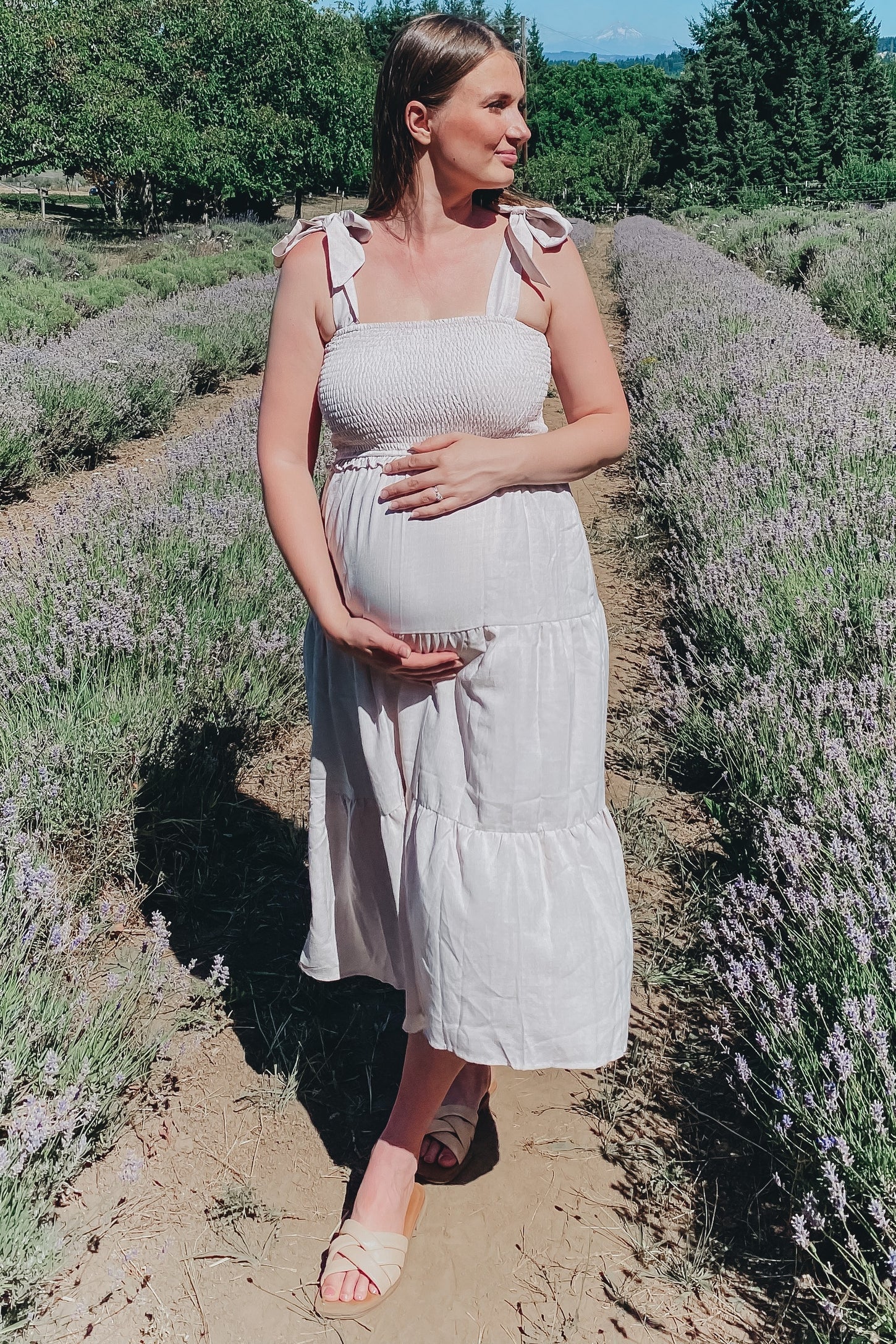 woman wearing a white smocked maternity dress with tie straps and a visible baby bump standing in a lavender field