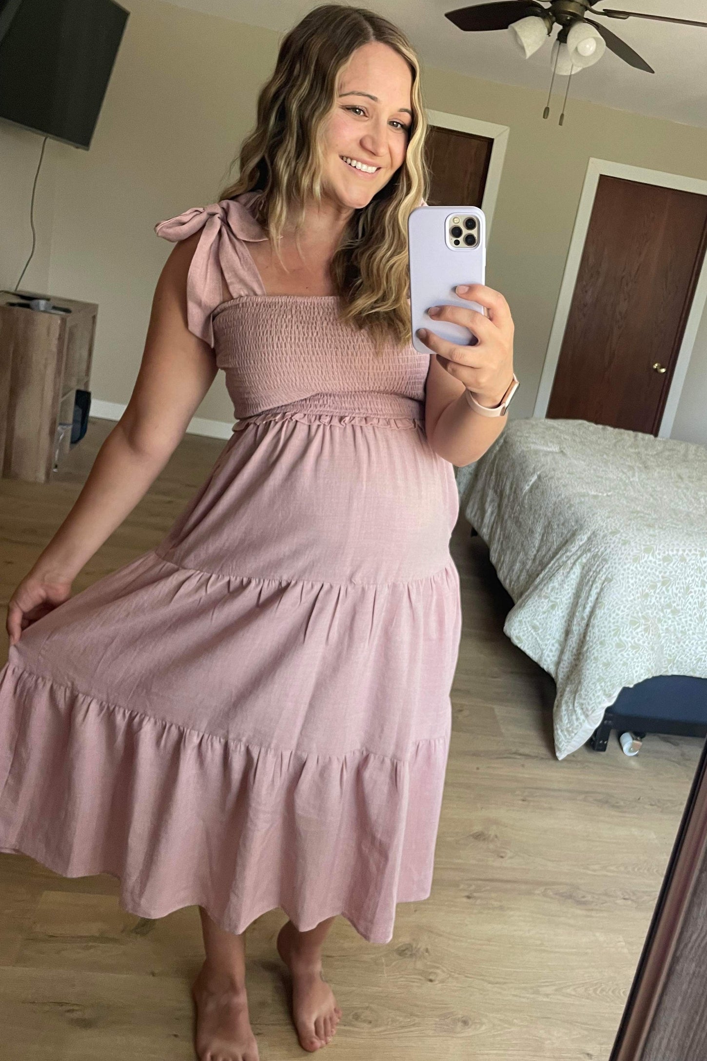 woman wearing a pink maternity dress with smocked bodice and tie straps, showing her baby bump while smiling in a mirror selfie