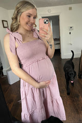 woman wearing a pink maternity dress with tie straps and a tiered skirt, holding her baby bump in front of a mirror
