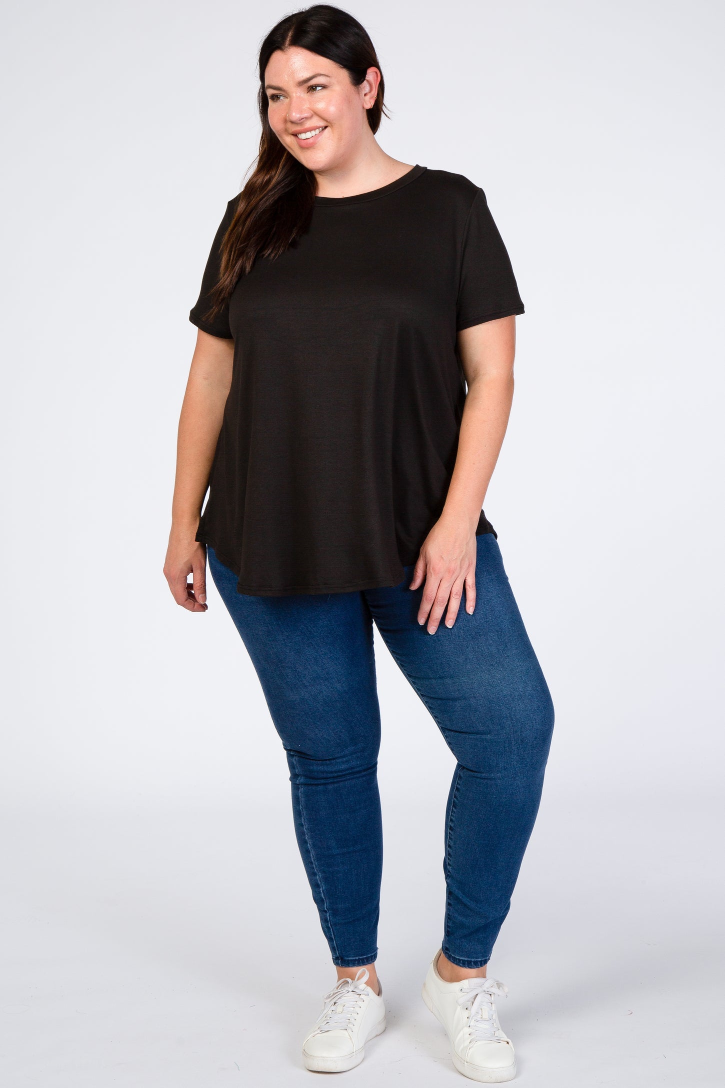 smiling woman wearing a black short sleeve tunic top and blue skinny jeans with white sneakers