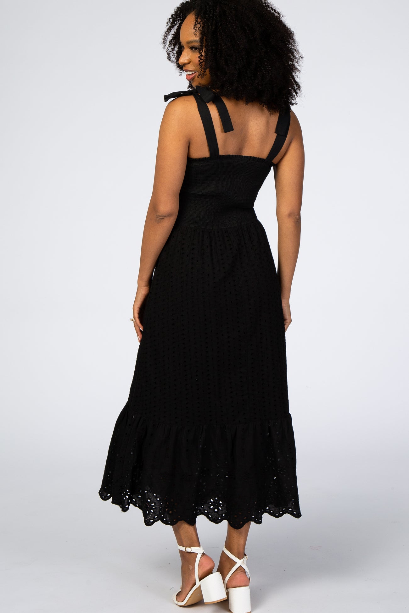 Woman in a black eyelet midi dress with tie straps and a ruffled hem, viewed from the back against a white background