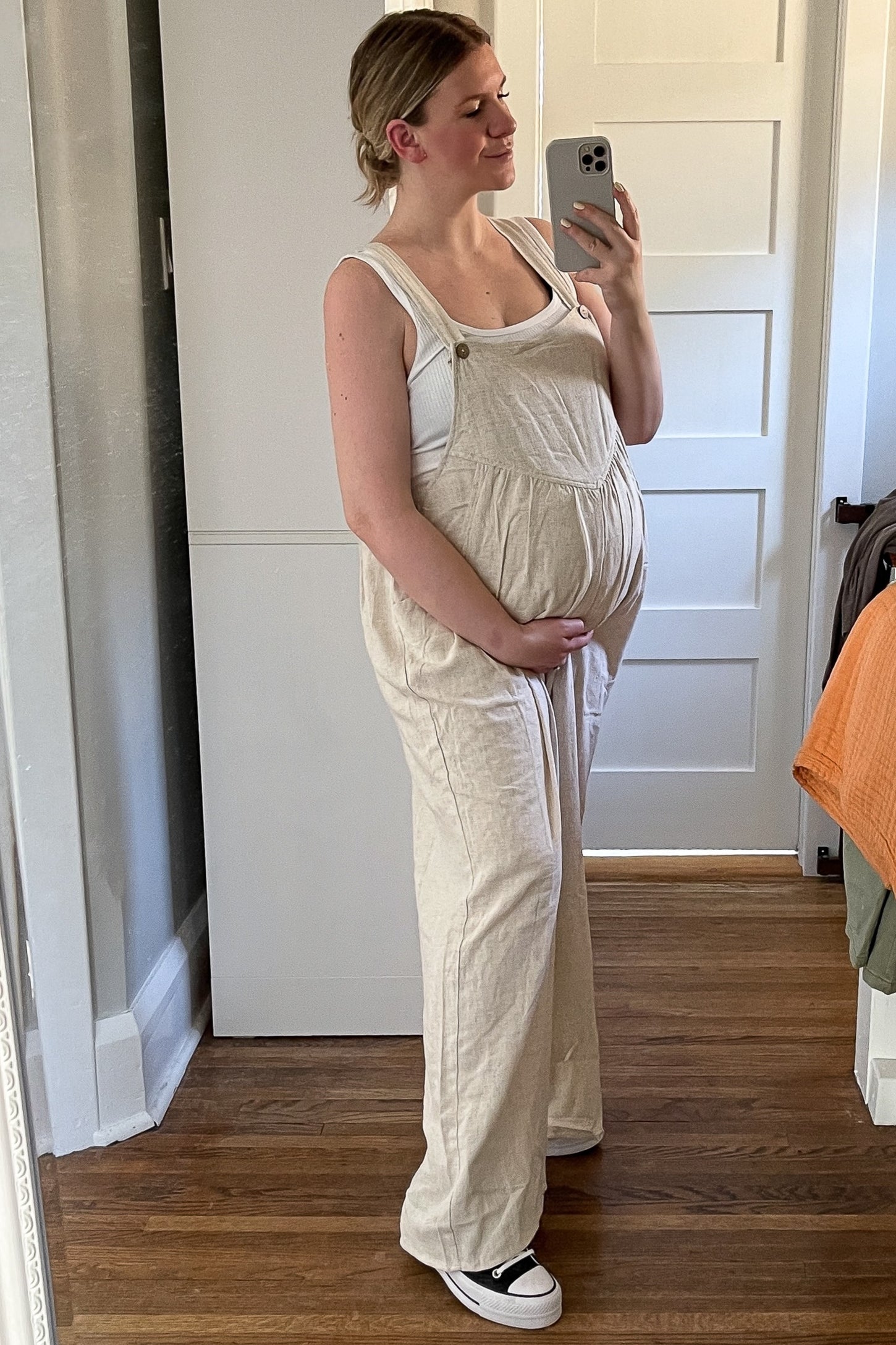 pregnant woman in beige maternity overalls and white tank top taking a mirror selfie