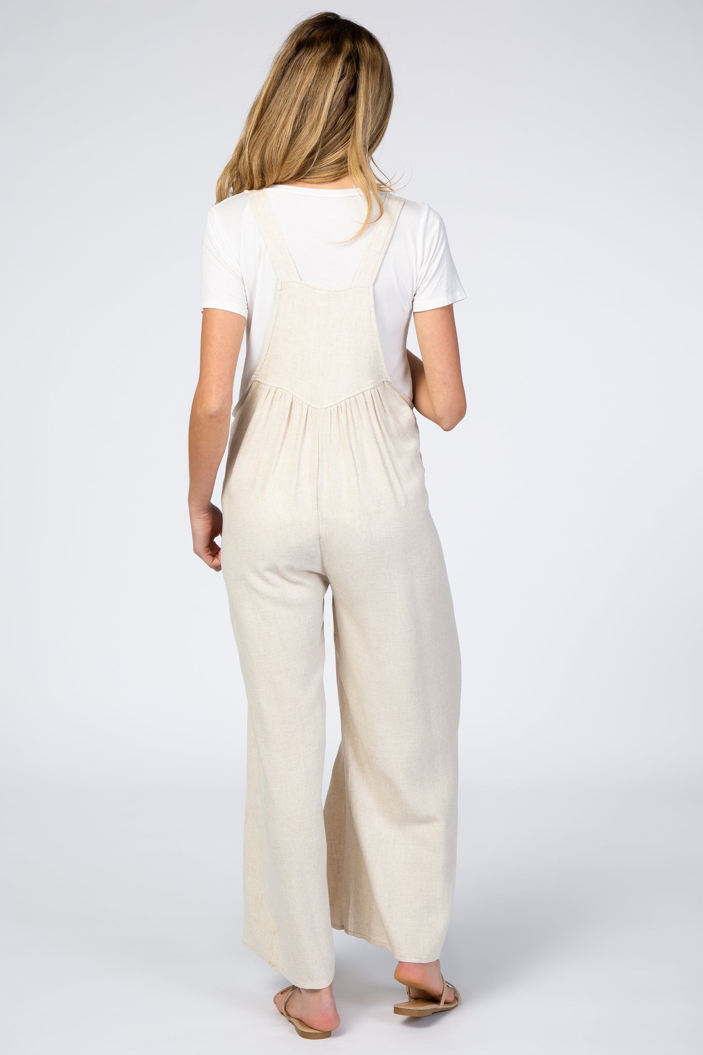 Back view of a woman wearing beige linen overalls over a white t-shirt with wide leg pants