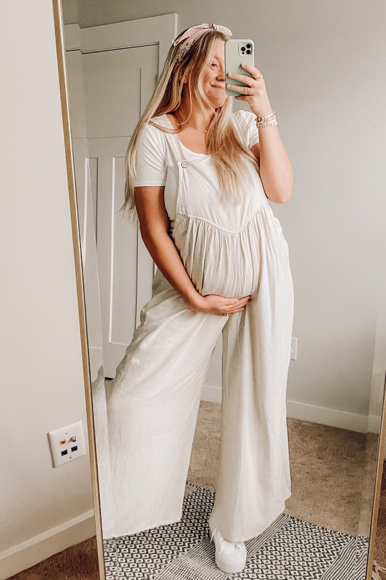 pregnant woman in white maternity overalls taking a mirror selfie while holding her baby bump