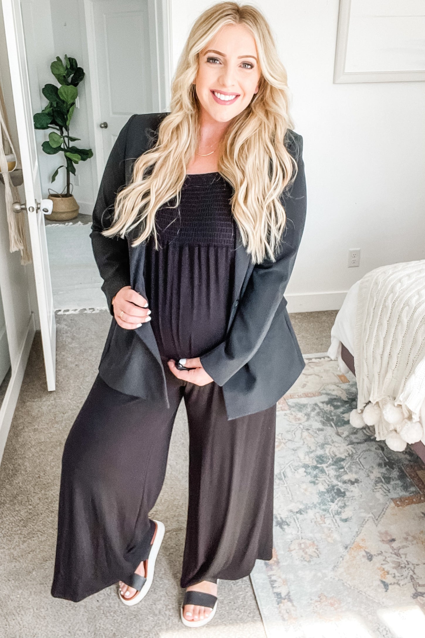 Smiling pregnant woman wearing a black blazer and jumpsuit standing in a bright bedroom