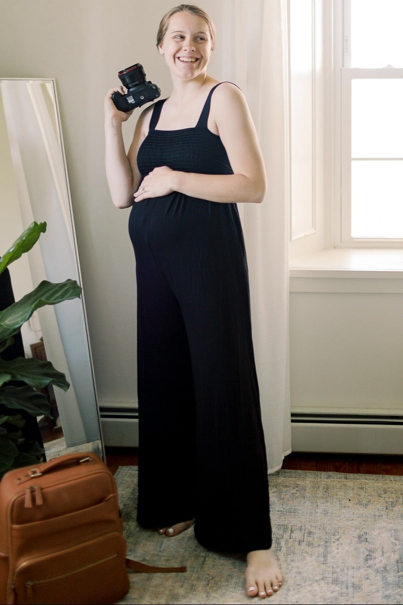 woman wearing a black maternity jumpsuit holding a camera and showing her baby bump