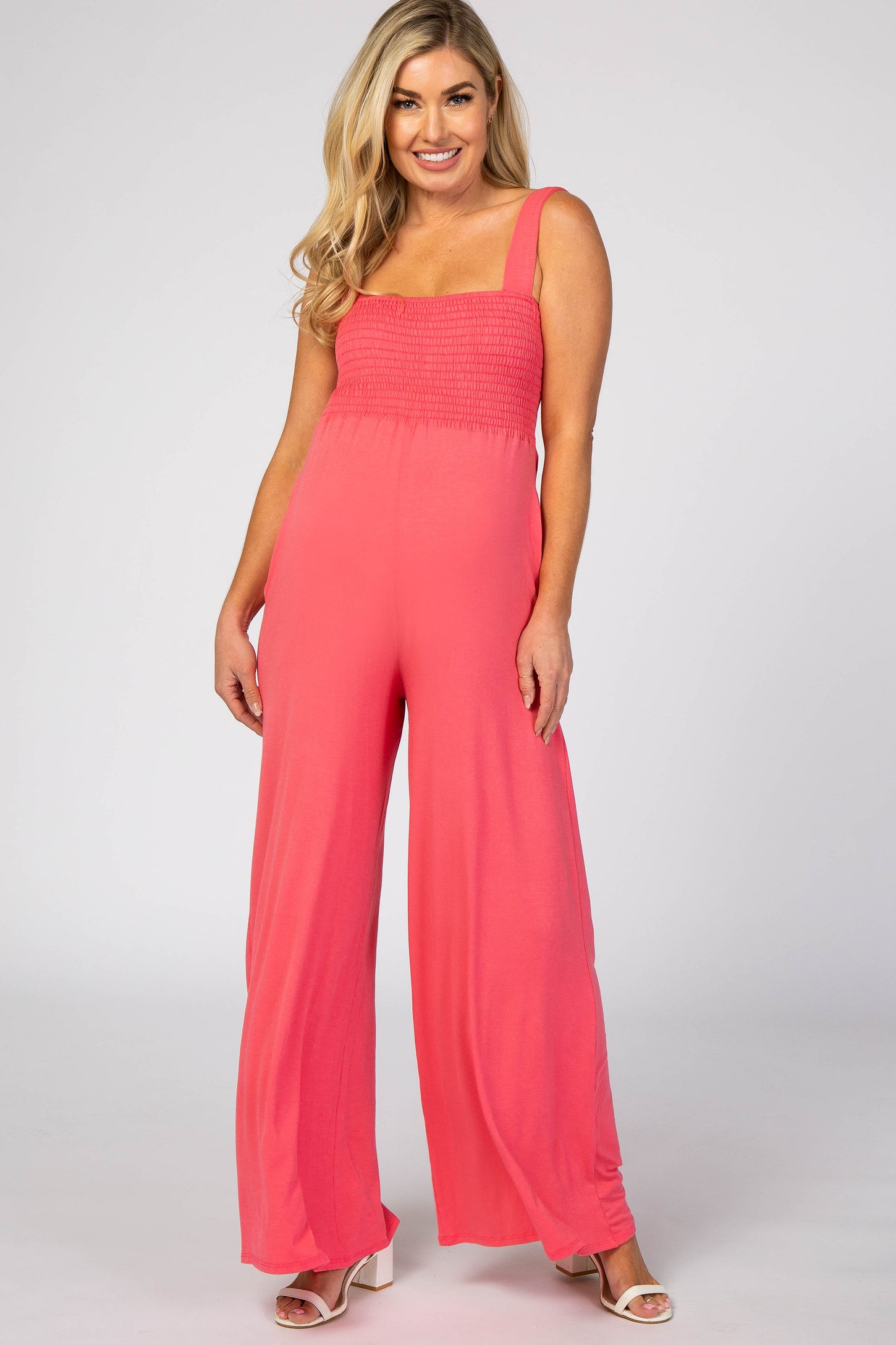 woman wearing a coral pink maternity jumpsuit with smocked bodice and wide-legged pants, model poses smiling