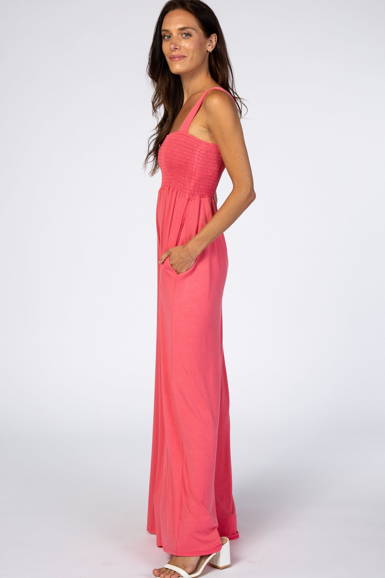 woman wearing a coral sleeveless maxi dress with a smocked bodice and pockets, standing sideways against a white background