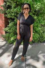 woman wearing a black short sleeve jumpsuit with a tie waist and brown loafers standing in front of green bushes