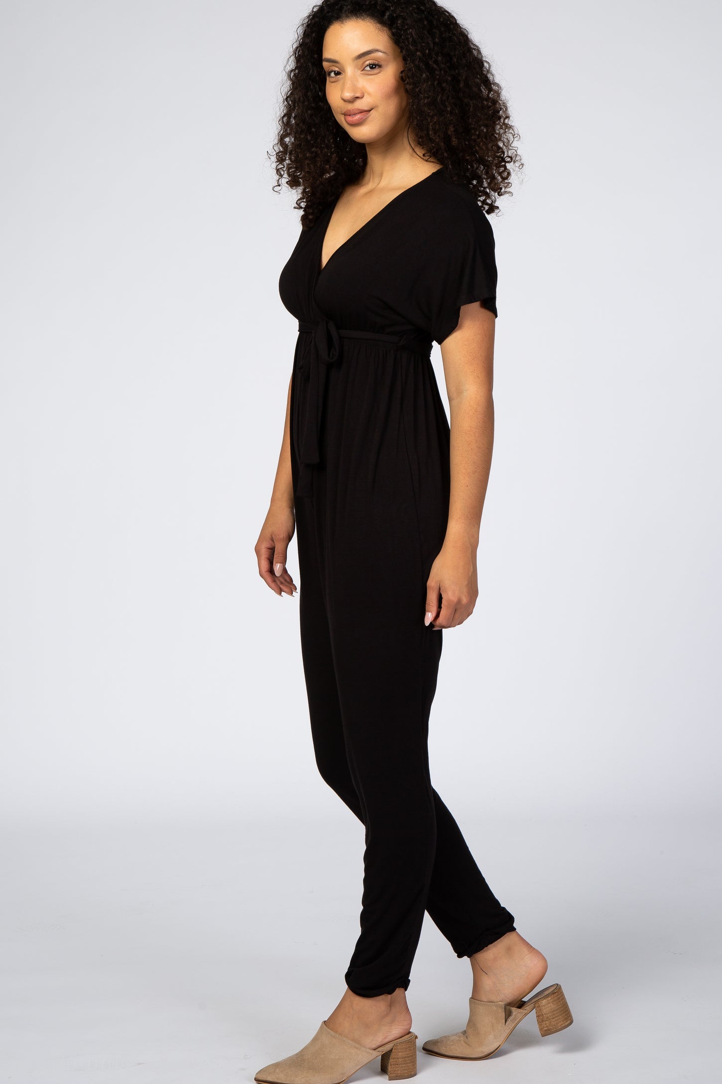 woman wearing a black short sleeve jumpsuit with a tie waist, standing sideways with curly hair and tan shoes