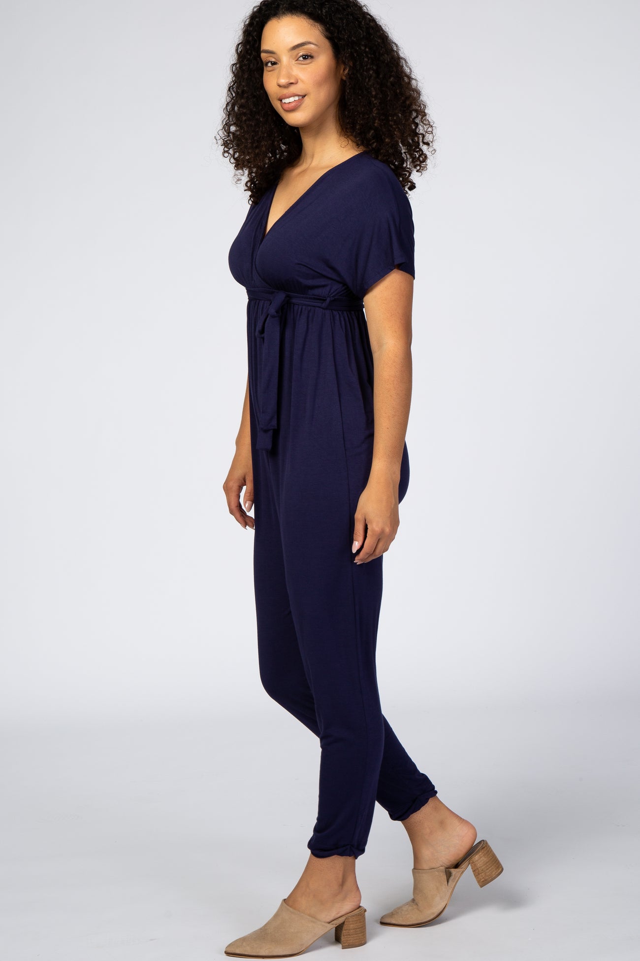 woman with curly hair wearing a navy blue short sleeve maternity jumpsuit and tan mule heels