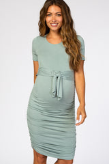 smiling pregnant woman wearing a sage green short sleeve maternity dress with a front tie and ruched sides