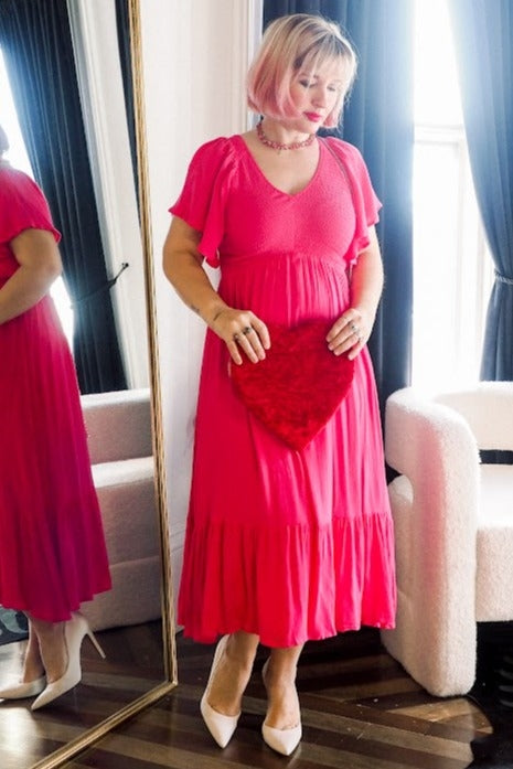 woman wearing a vibrant pink midi dress with short flutter sleeves and a tiered hem, holding a heart-shaped purse