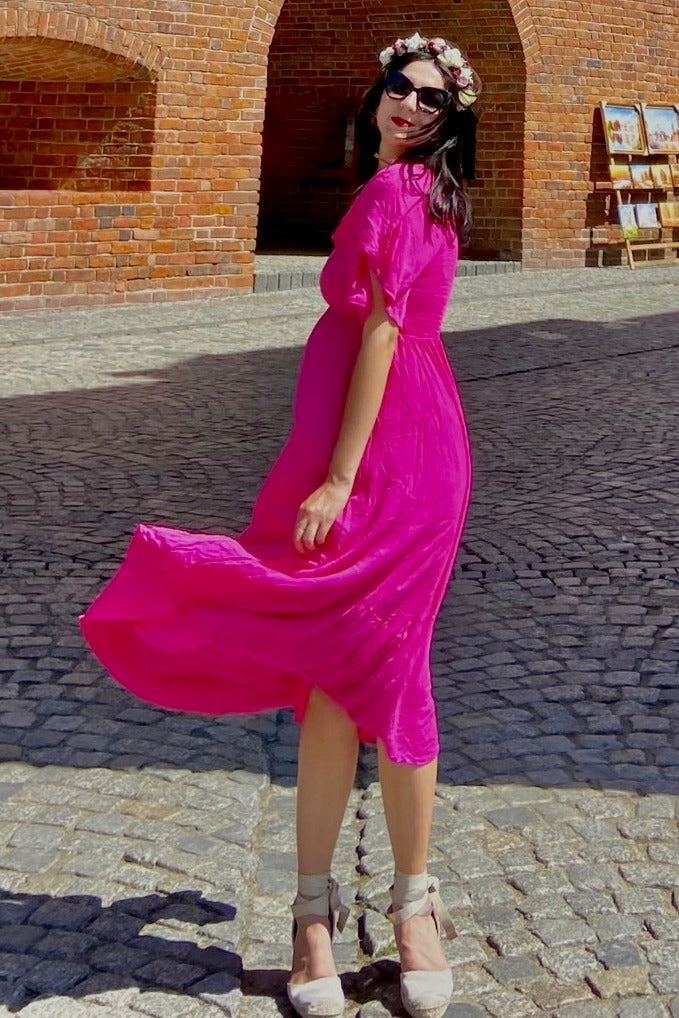 woman wearing a bright pink short sleeve dress, twirling in a cobblestone area with sunglasses and a floral headband