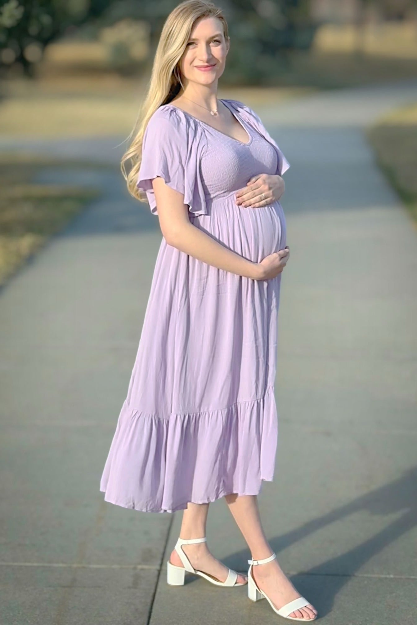 woman wearing a light purple maternity dress with flutter sleeves and a gathered waist, holding her baby bump