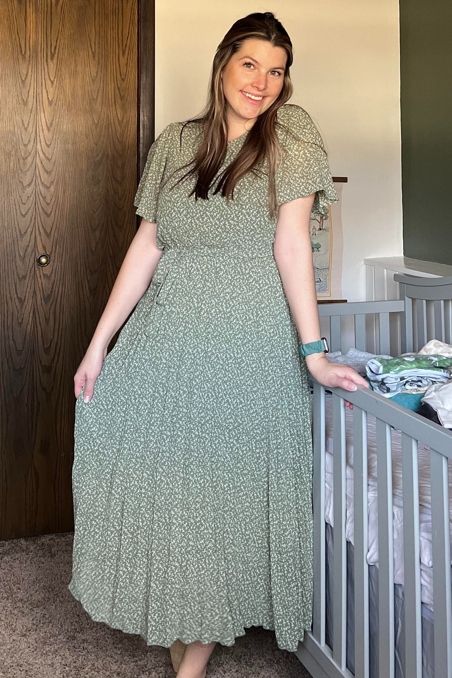 woman wearing a green floral maxi dress with flutter sleeves and a fitted waist, standing next to a crib