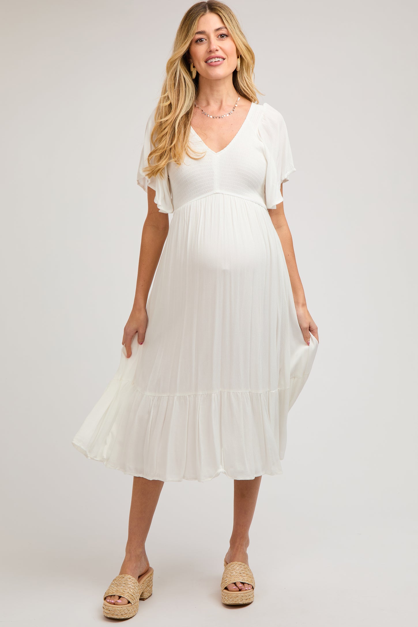 woman wearing a white maternity dress with a v-neck and flutter sleeves, holding her baby bump