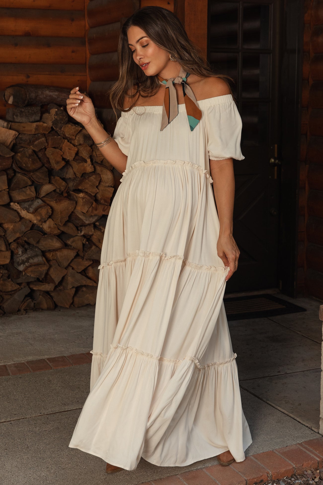 woman wearing a cream off-shoulder maternity maxi dress with tiered ruffles and a decorative neck scarf