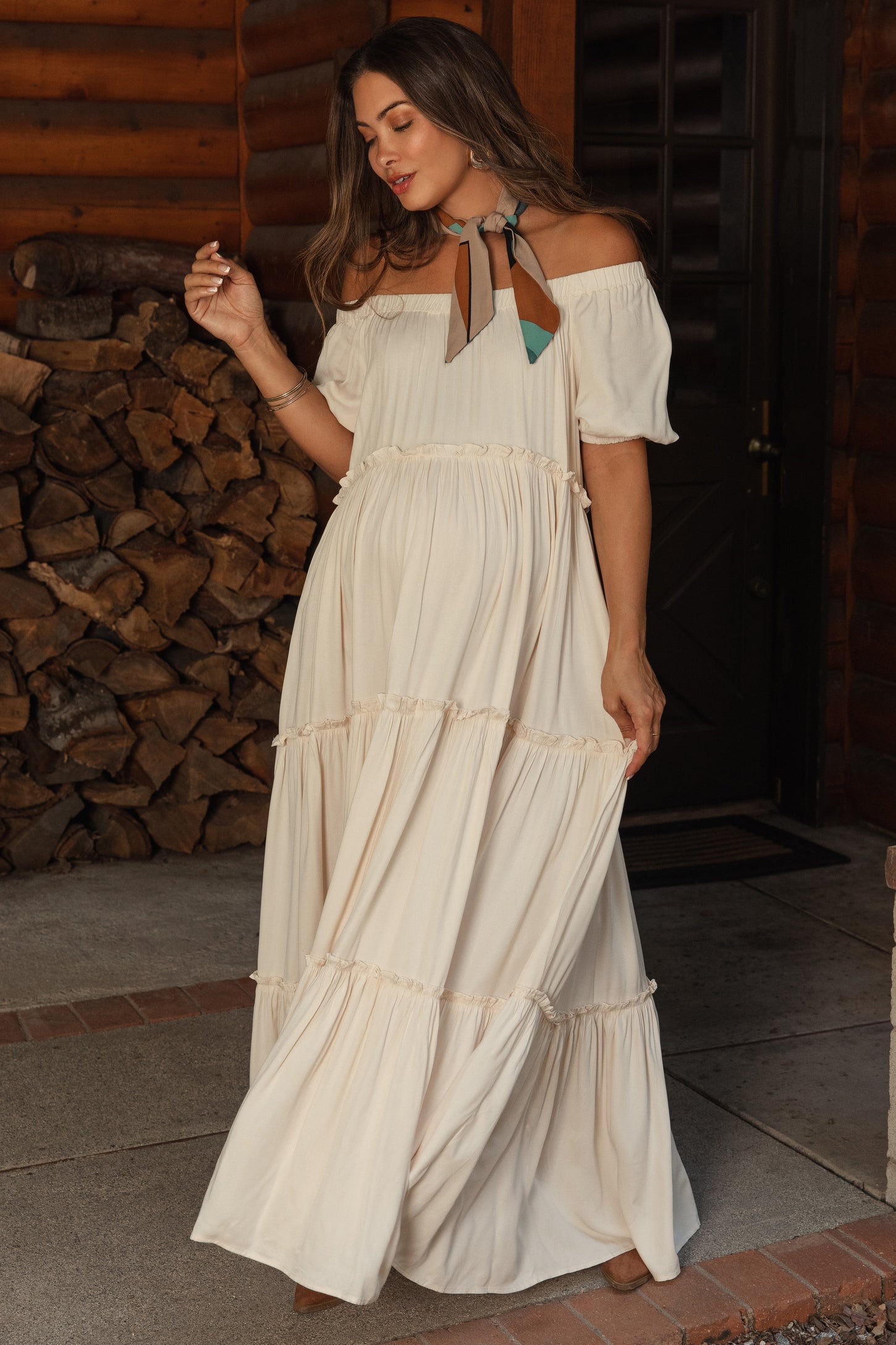 woman wearing a cream off-shoulder maternity maxi dress with tiered ruffles and a decorative neck scarf