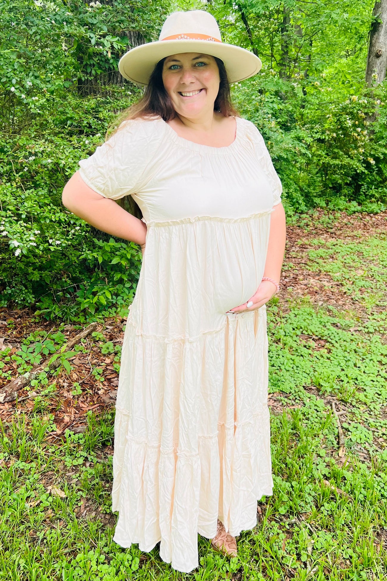 woman wearing a cream maternity maxi dress with tiered details, holding her baby bump in a natural outdoor setting