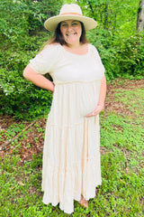 woman wearing a cream maternity maxi dress with tiered details, holding her baby bump in a natural outdoor setting
